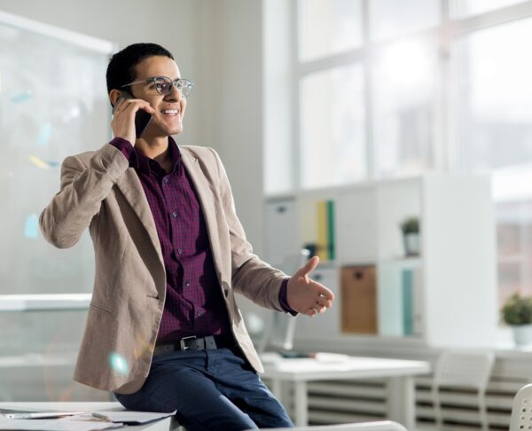 Successful broker in elegant formalwear sitting on desk and consulting one of his clients by the phone